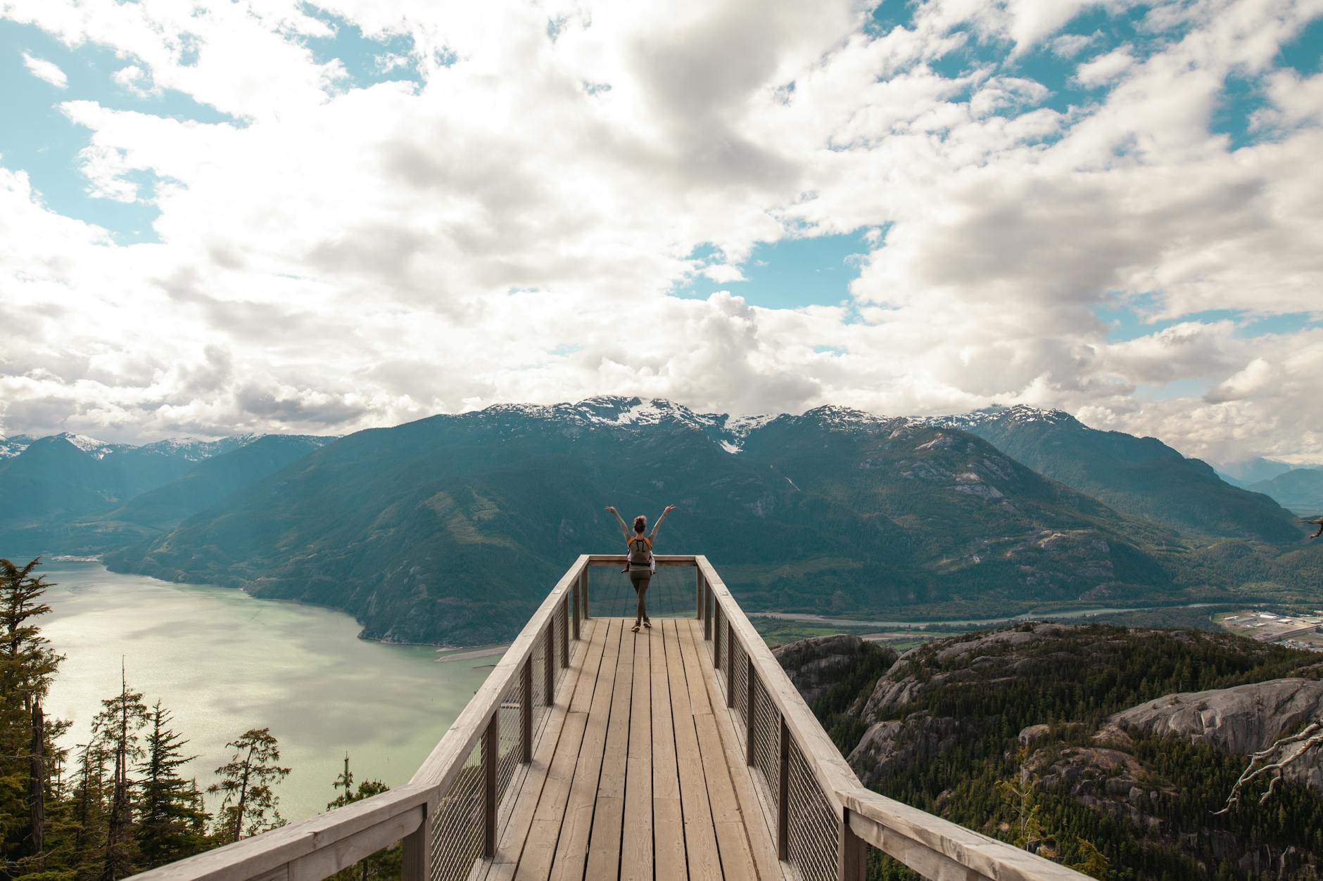 person standing with hands on air on brown wooden dock with overlooking view of lake under white clouds and blue sky